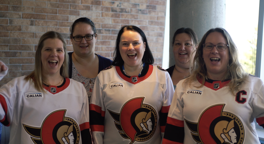 Five women stand indoors, smiling at the camera; three of them wear Ottawa Senators hockey jerseys, and a brick wall is visible in the background.